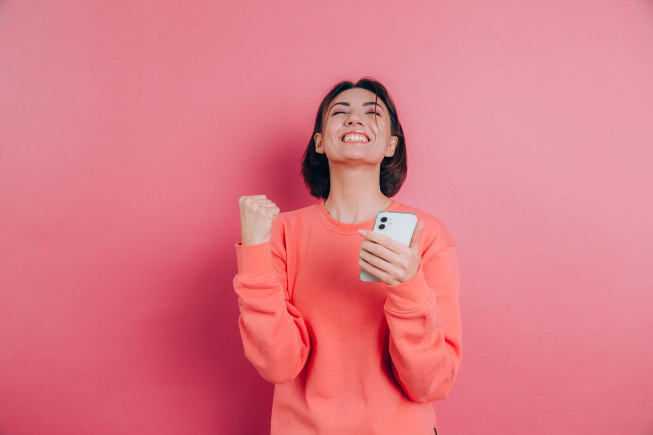 Young woman in orange pullover against pink background clenches a fist in joy and she throws her bead back in victory, holding her smartphone.