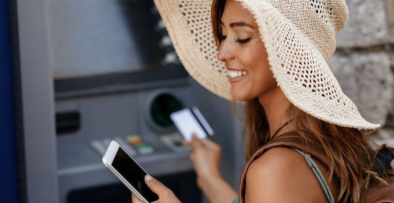 Young happy woman withdrawing money from cash machine while using smart phone after learning, "How to put money on Cash App card at ATM."
