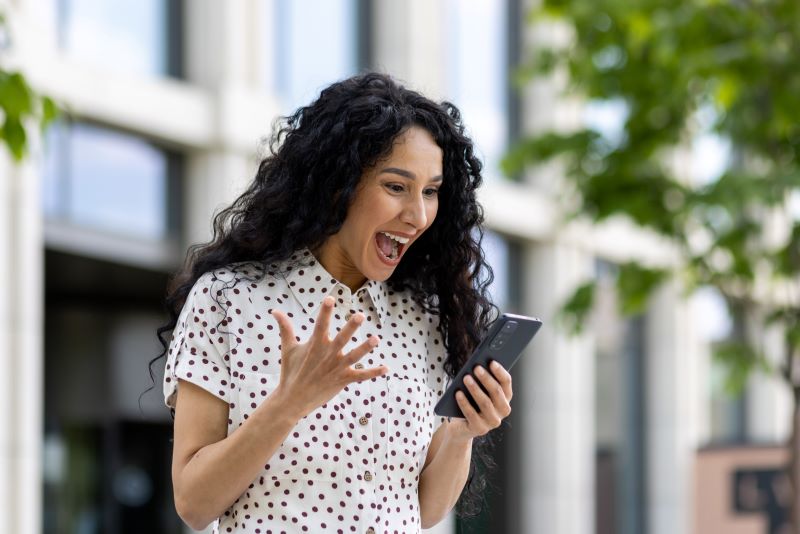 Woman outdoors looking at phone with excitement and raised hand.