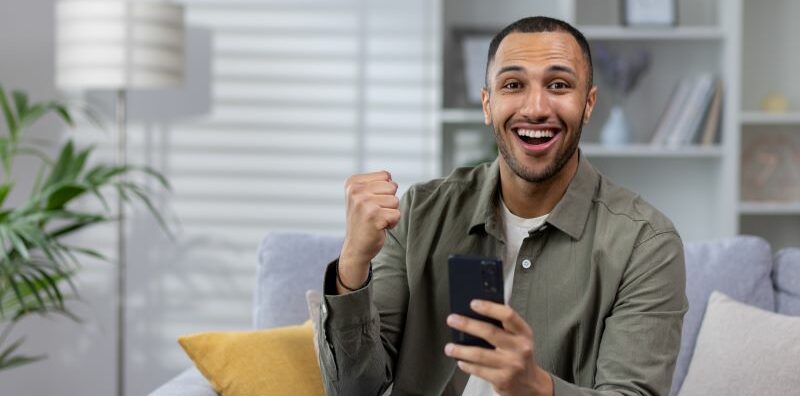 Man on couch holding cell phone, raising hand, and smiling.