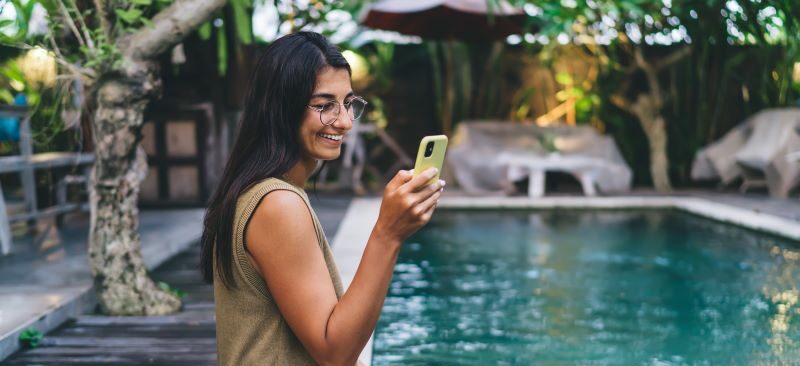 Woman outside sitting with feet in pool smiling at cell phone