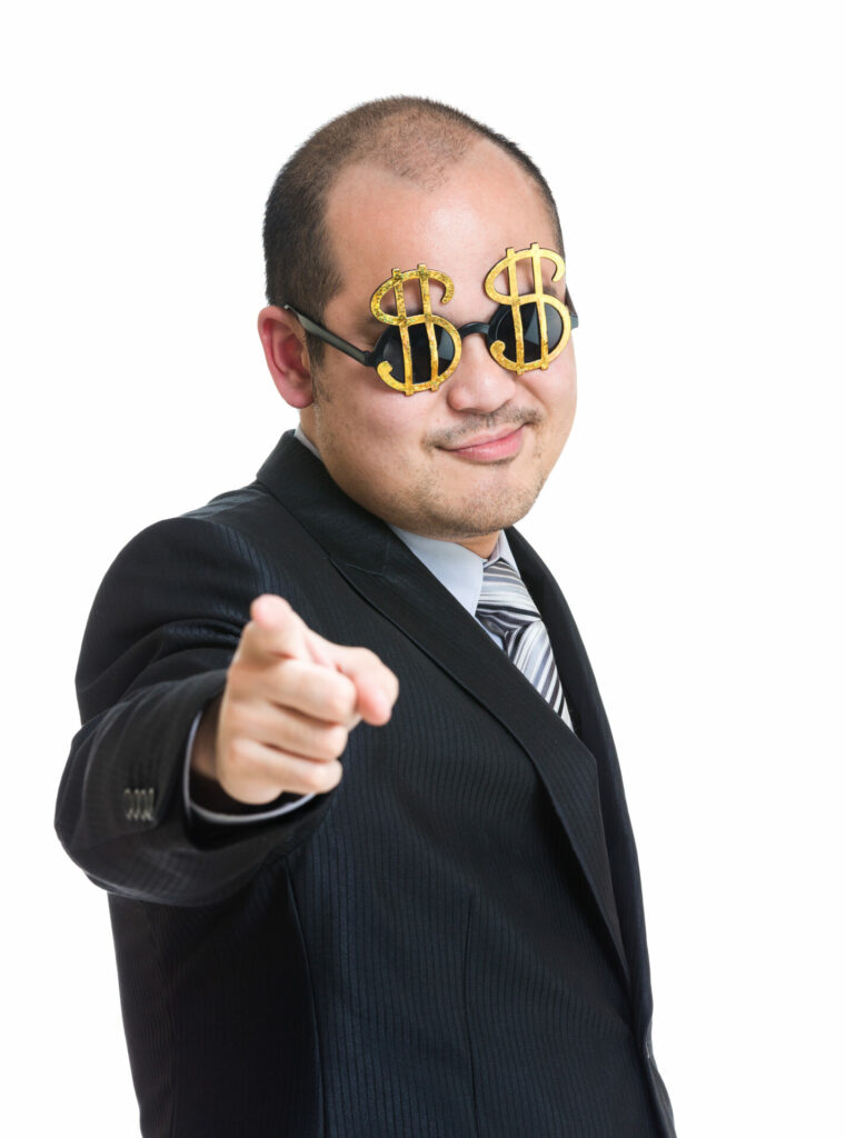 Smug man in suit and dark glasses decorated in two large gold dollar bills points at camera in a winning way.