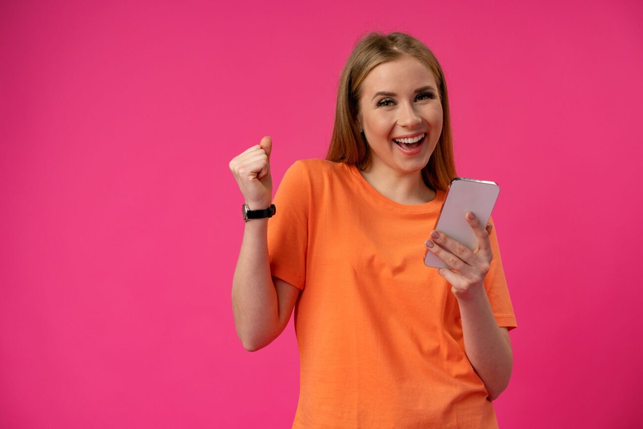 Young woman in orange T-shirt against pink background smiles while holding her smartphone in one hand and raising the other in victory.