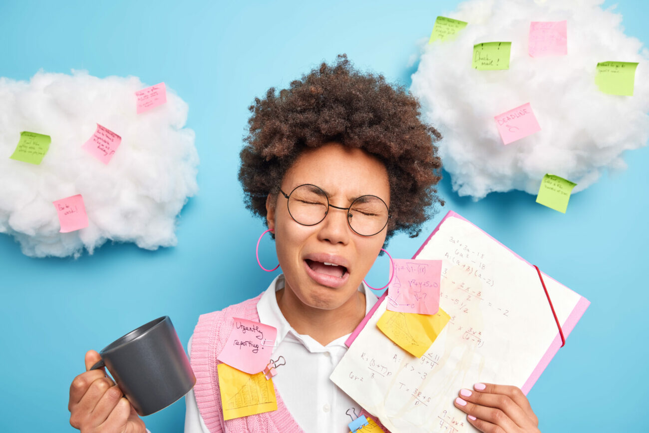 Young woman play-weeps from stress as clouds filled with Post-It notes surround her head.