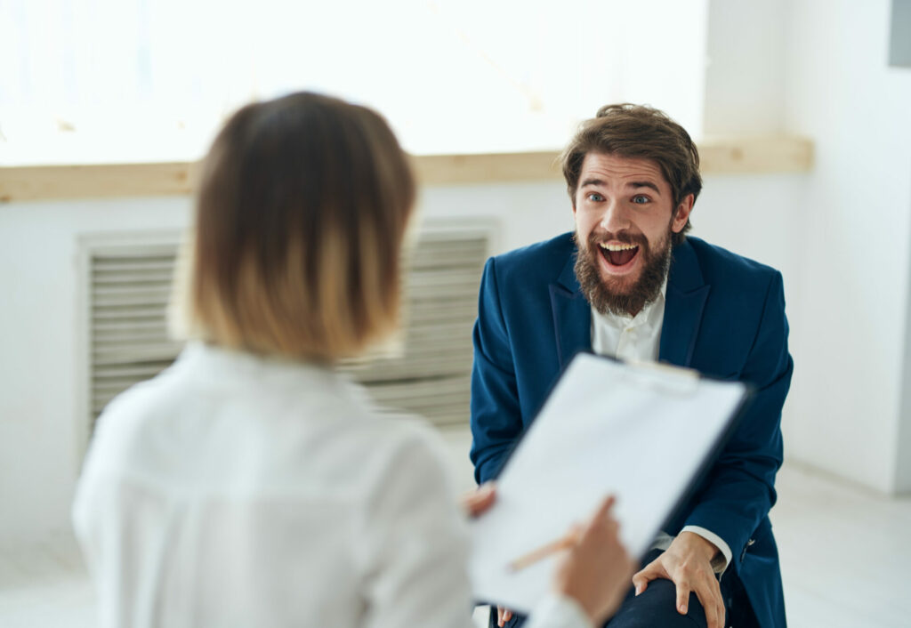 Overtly happy man with beard and blue suit jacket laughs in an exaggerated way while grasping his knees as he is interviewed by a woman focused on her survey clipboard.