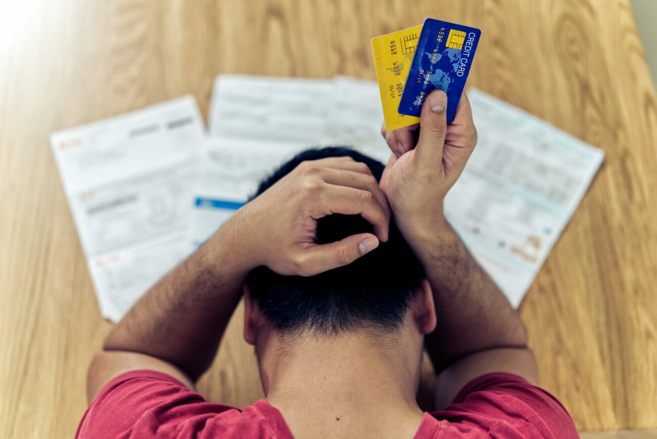 Top view of stressed young Asian man holding credit cards and thinking about finding money to pay credit card debt and all bills. He is holding head by another hand. Financial problem concept.