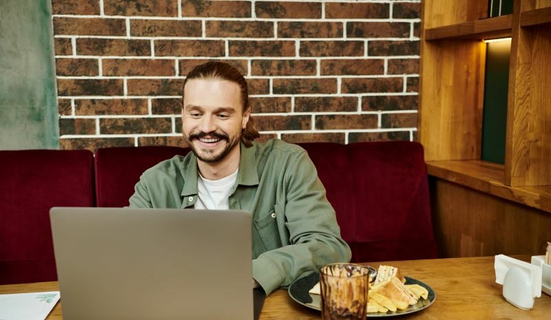 Man smiling at laptop while sitting at table in front of brick wall
