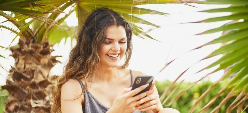 Woman sitting under palm tree smiling at cell phone