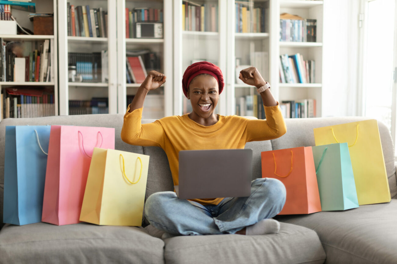Emotional cheerful african american female consumer purchasing from home, young black lady sitting on couch among colorful shopping bags, using laptop, raising hands up.