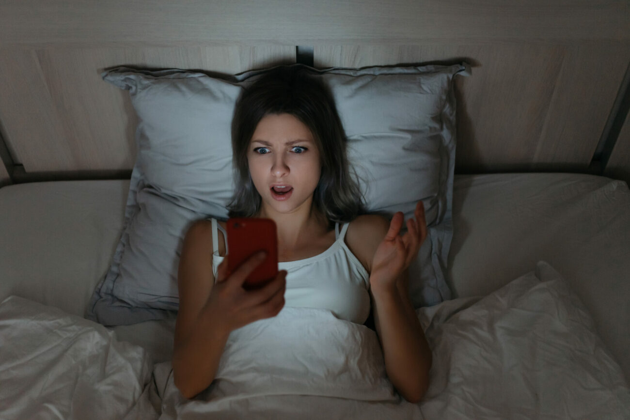 Woman lays in bed in room lit by the smartphone she is staring in surprise at.