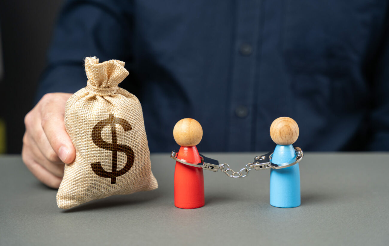 A sack with a dollar sign on it sits on a table with two figurines that are chained together, representing debt.