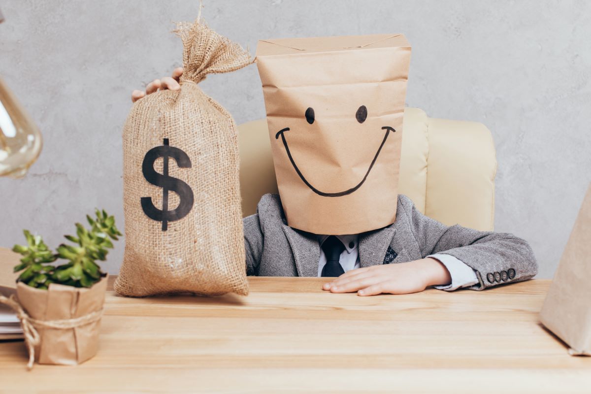 A young man with a smiley face on a bag that is over his head sits at a desk beside a large sack with a dollar sign printed on it.