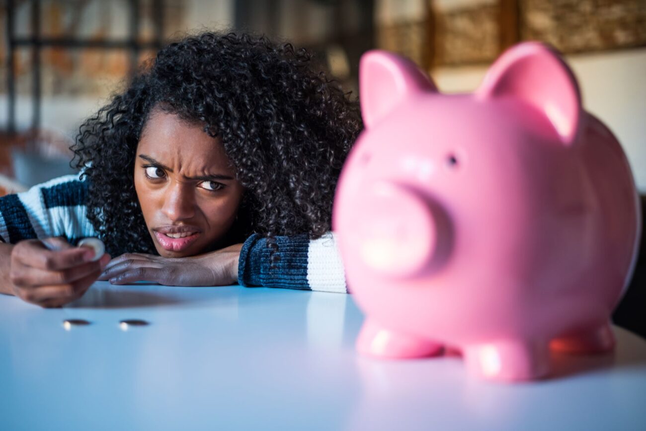 Young black woman rests her chin on here hands upon a desk with some small change on it as she looks pensively at the pink piggy bank standing nearby.