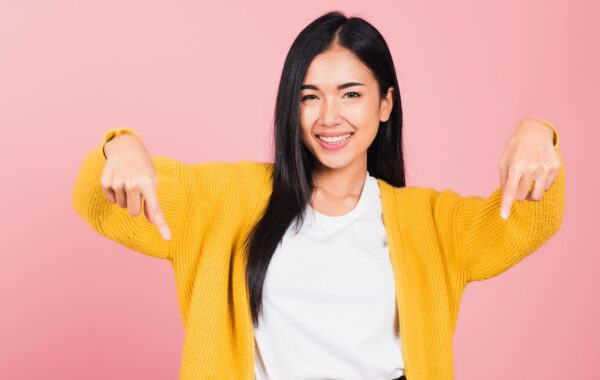 Young Asian woman in yellow cardigan smiles as she raises her arms and points directly downwards.