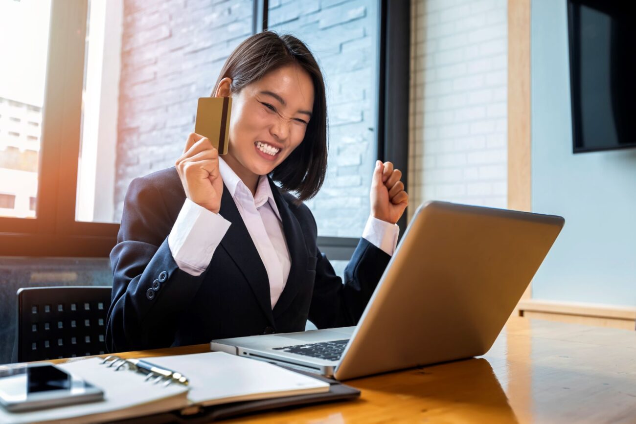 Happy Asian business-woman sitting at desk in suit and holding a credit card, throws up her hands and smiles winningly.
