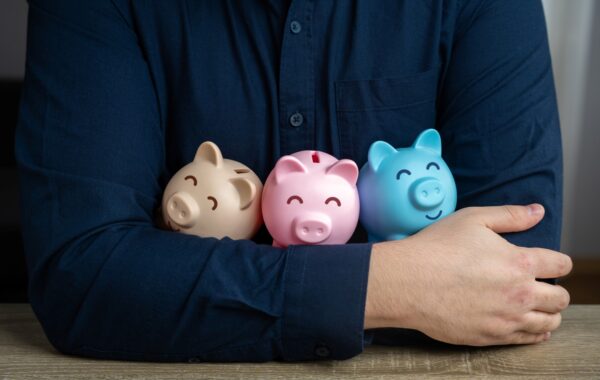 Close up image of three small piggy banks, one brown, one pink and one light blue being held in the arms of an unseen male wearing a dark blue shirt.