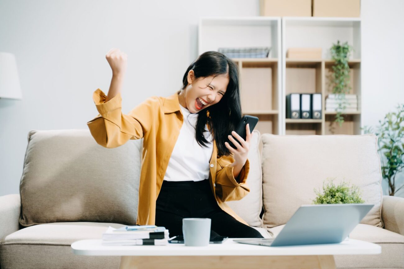 Young Asian woman sitting at a makeshift desk or coffee table at home looks into her smartphone and throws her free hand up with joy.