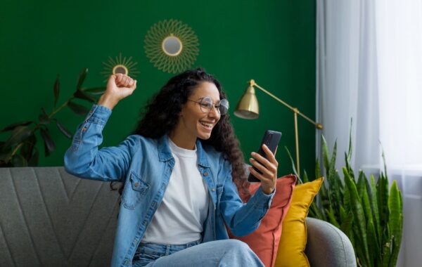 Young Hispanic woman at home holds her smartphone in one hand and raises the other in victory and she smiles.