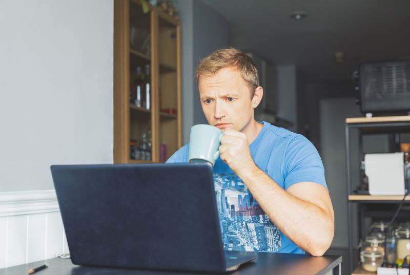 Man drinking coffee at home looking curiously at computer screen