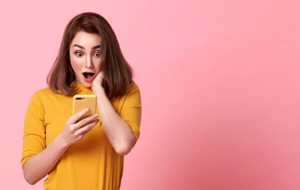 Young woman against pink background gasps in surprise as she looks at her smart phone.
