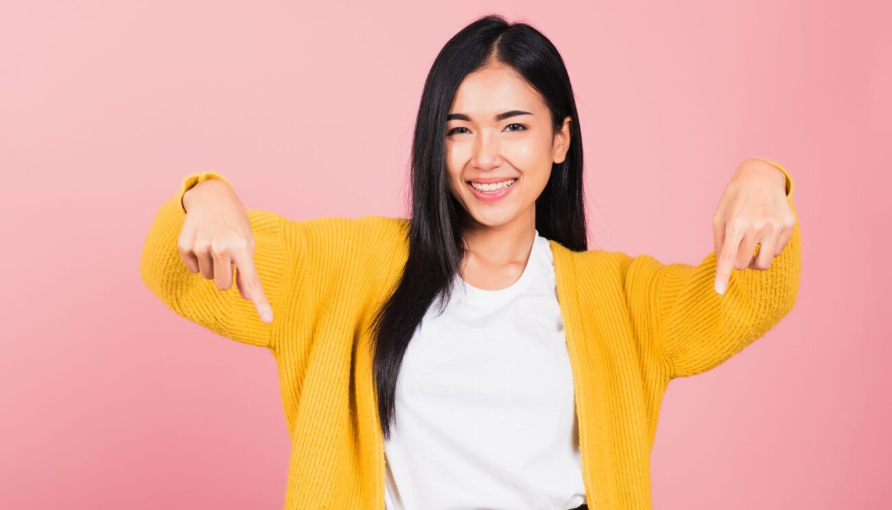 Young Asian woman against pink background and wearing a bright yellow cardigan smiles and points downwards.