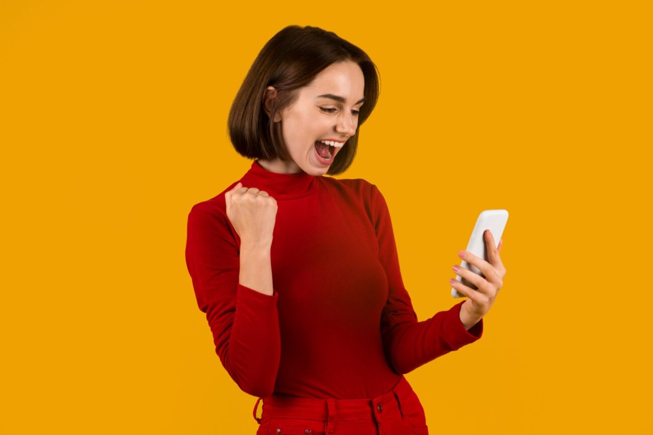 Young woman in casual red clothing against a yellow background shouts and raises an arm in victory and she looks at her smart phone.
