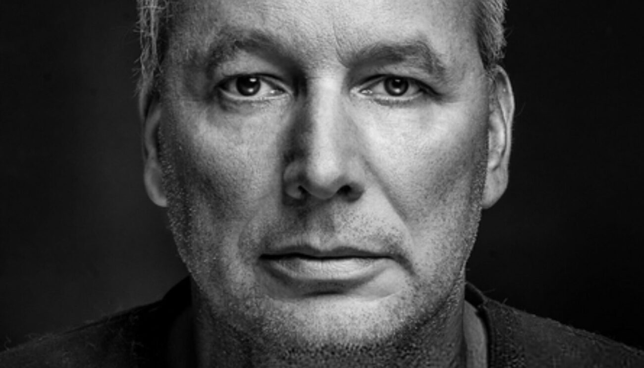 Close up of a solemn looking mature man with gray hair in black t-shirt against dark background.