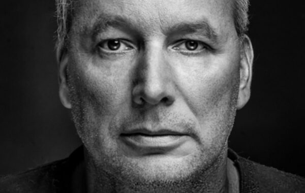 Close up of a solemn looking mature man with gray hair in black t-shirt against dark background.