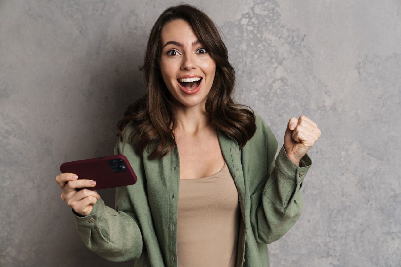 Young woman in green jacket against gray background laughs in delight after success on her smart phone.