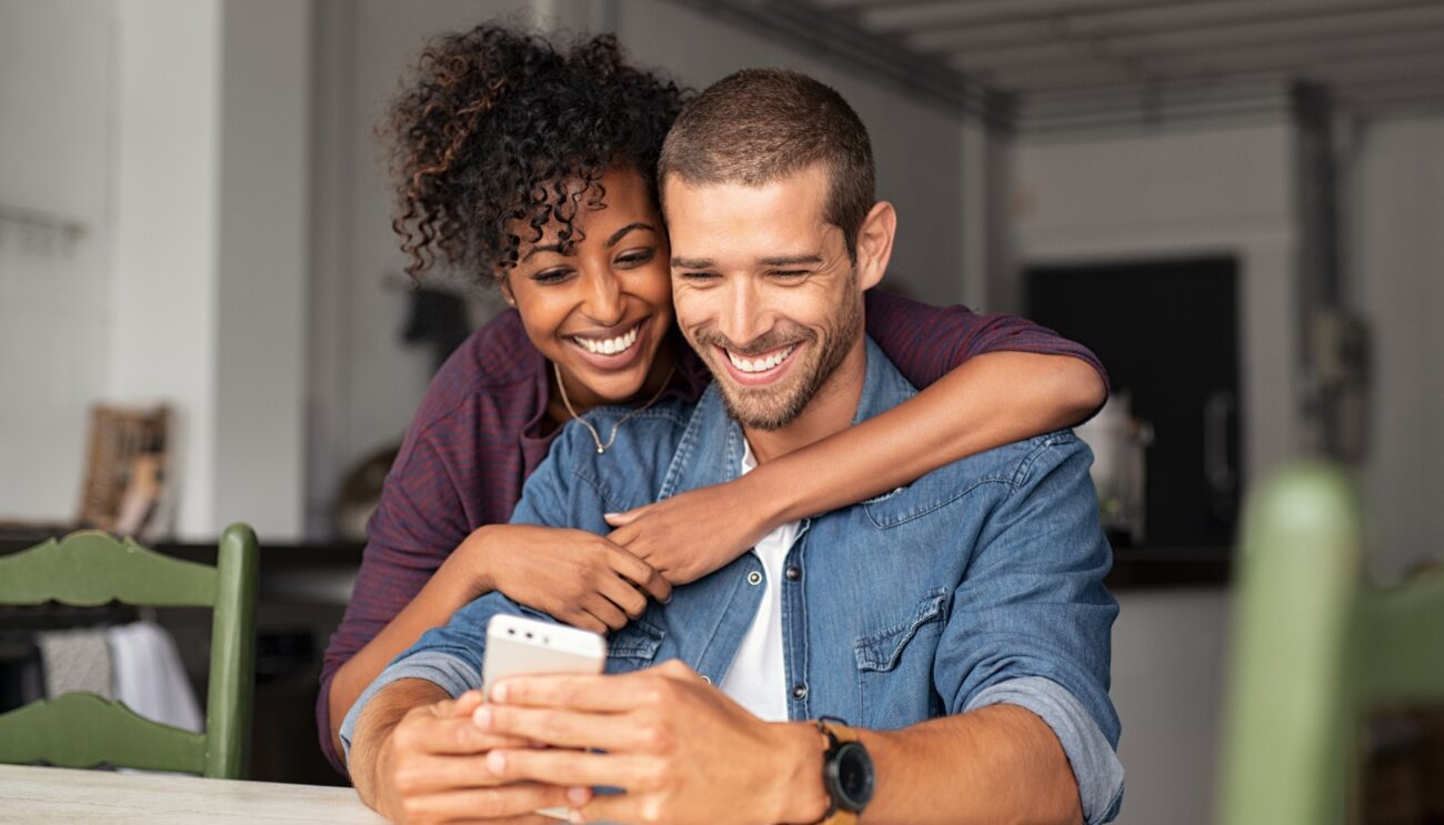 Happy couple smile and hug as they view something on a cellphone.