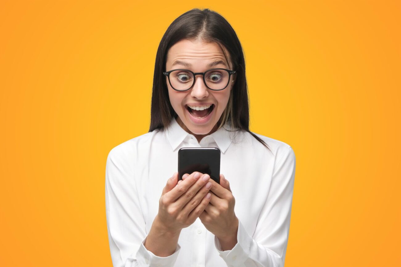 Young lady in spectacles against yellow background looks happily surprised as she looks at her smartphone.