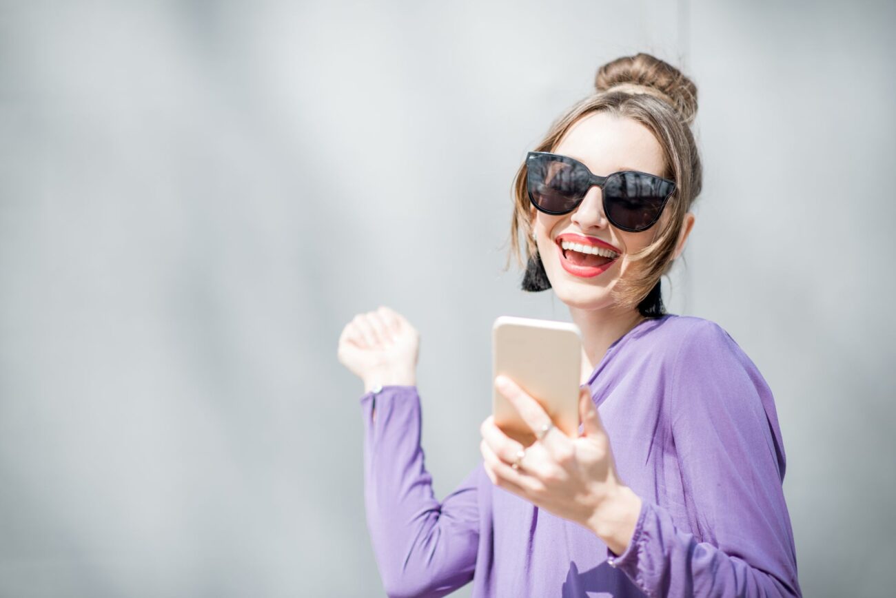 Joyous woman wearing large sunglasses outside throws up a victory fist after reading something on here smartphone.