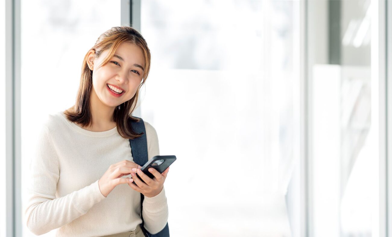 Young brunette woman with backpack smiles as she looks up from her smart phone.