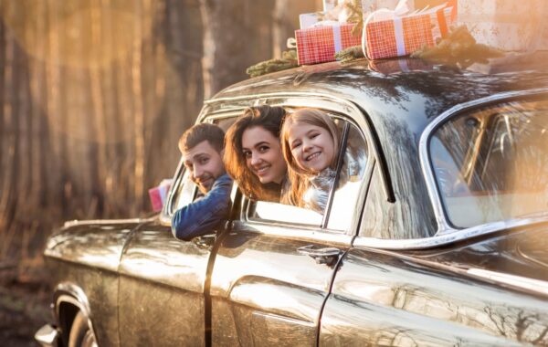 Happy couple and daughter leaning out of stationary car windows, on top of which are gift-wrapped gifts.
