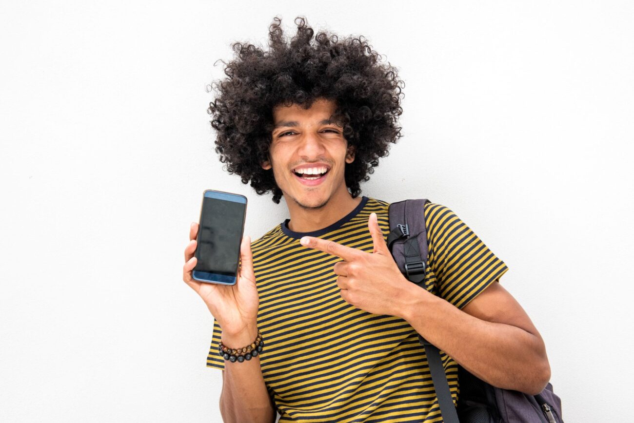 Happy student with backpack smiles at viewer and points to smartphone.