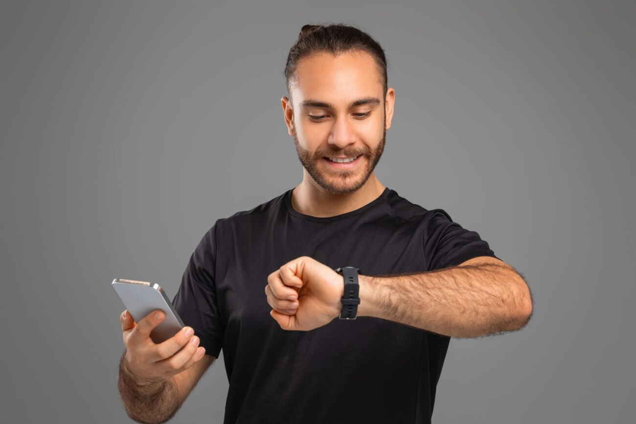Young man checks his watch and smiles while holding his smartphone.