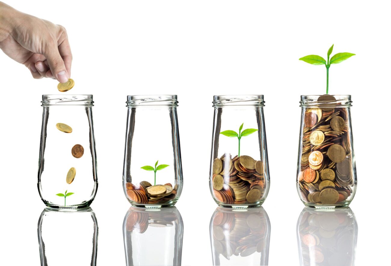 Four glass jars in a row contain pennies. The jar on the left is starting to be filled, while the jar on the right is full and shows a plant growing from the top.