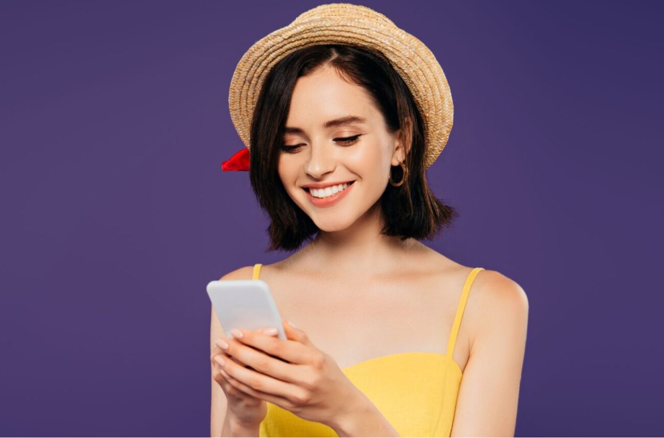 Smiling young woman in yellow dress and straw hat against purple background smiles into smartphone.