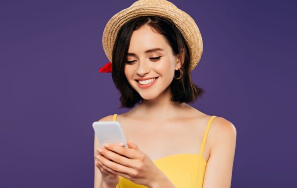 Smiling young woman in yellow dress and straw hat against purple background smiles into smartphone.